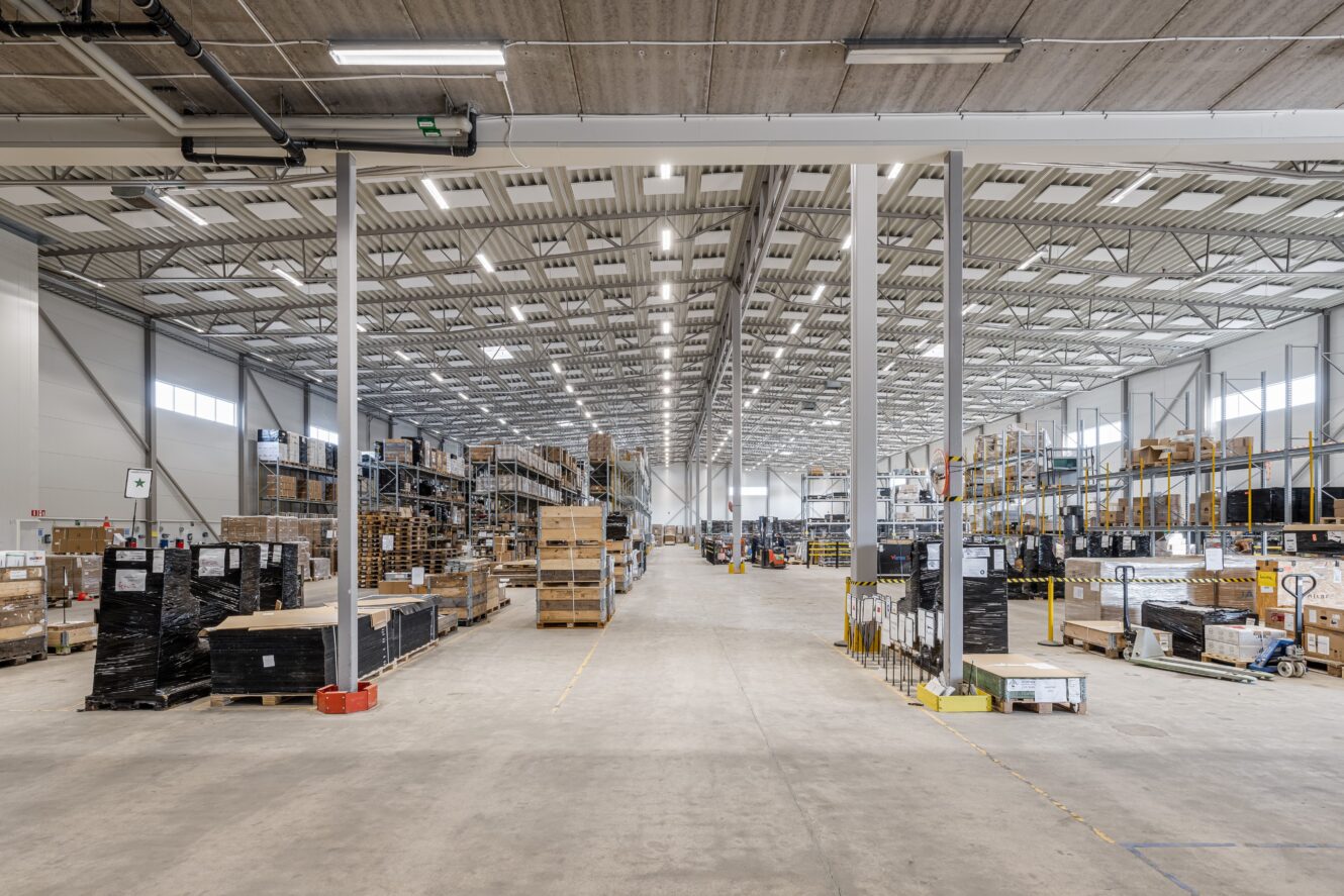 Bred vy över ett rymligt lager med höga hyllor, staplade pallar och olika lådor och utrustning som är organiserade längs båda sidor av en central gång.