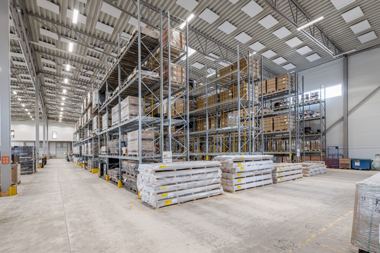 Stor lagerlokal med höga hyllor där pallar med varor förvaras, staplar med inslagna varor på golvet och stark takbelysning.