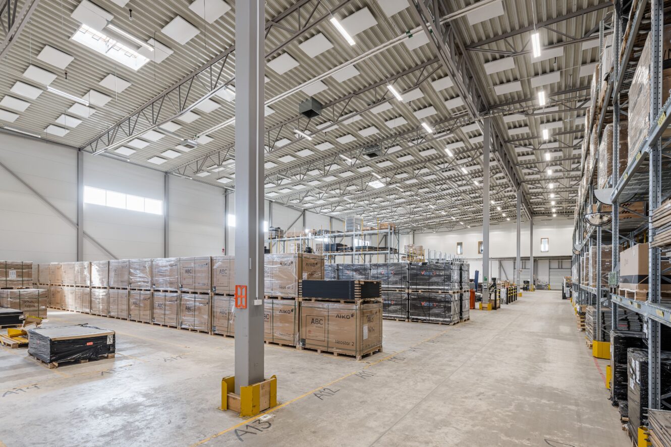 Stor lagerlokal med högt i tak, staplade pallar med lådvaror, metallhyllor och breda gångar under stark industribelysning.