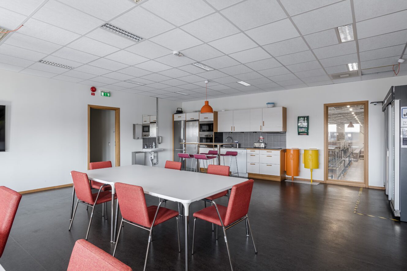 Ett modernt fikarum på ett kontor med ett vitt bord, röda stolar, pentry, kylskåp, orange och gula papperskorgar och stora fönster med utsikt över ett lagerområde.