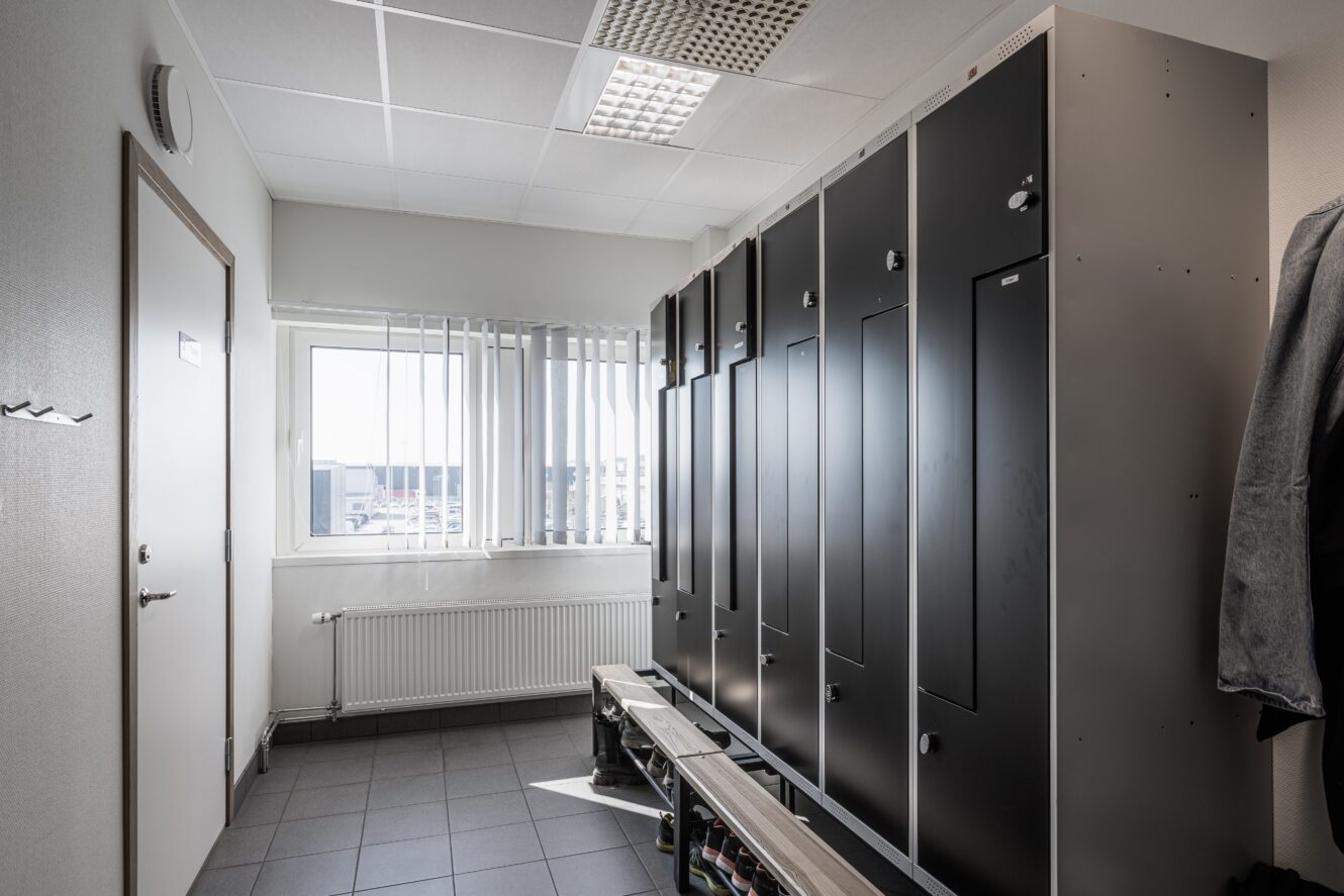 Ett omklädningsrum med svarta skåp, klinkergolv, ett fönster med vertikala persienner och en bänk i mitten. Under fönstret finns ett element och till höger hänger en kappa.