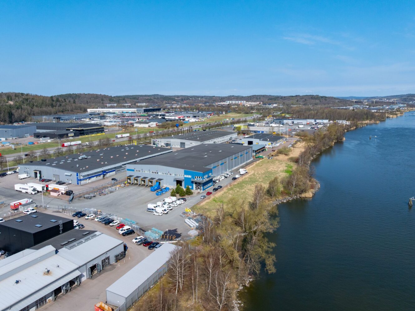 Flygfoto över ett industriområde med lagerbyggnader, parkerade fordon och en flod som rinner längs med anläggningen en klar dag.