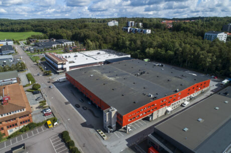 Flygfoto över en stor industrihall med röd fasad, omgiven av vägar, träd och närliggande kommersiella byggnader under en delvis molnig himmel.