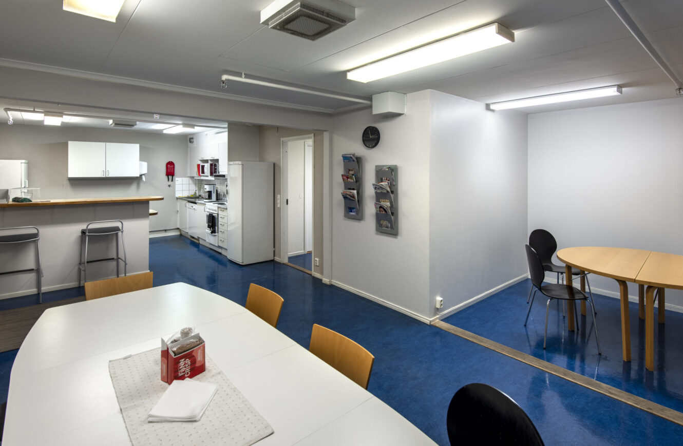 Ett fikarum för personalen med blått golv, vita väggar, bord och stolar, pentry med vitvaror och lysrör i taket.