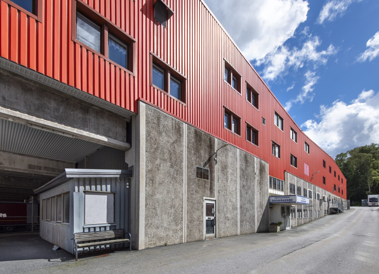 En stor industribyggnad med röd metallfasad och betongunderväggar, med flera fönster och småföretag i marknivå längs en sluttande väg.