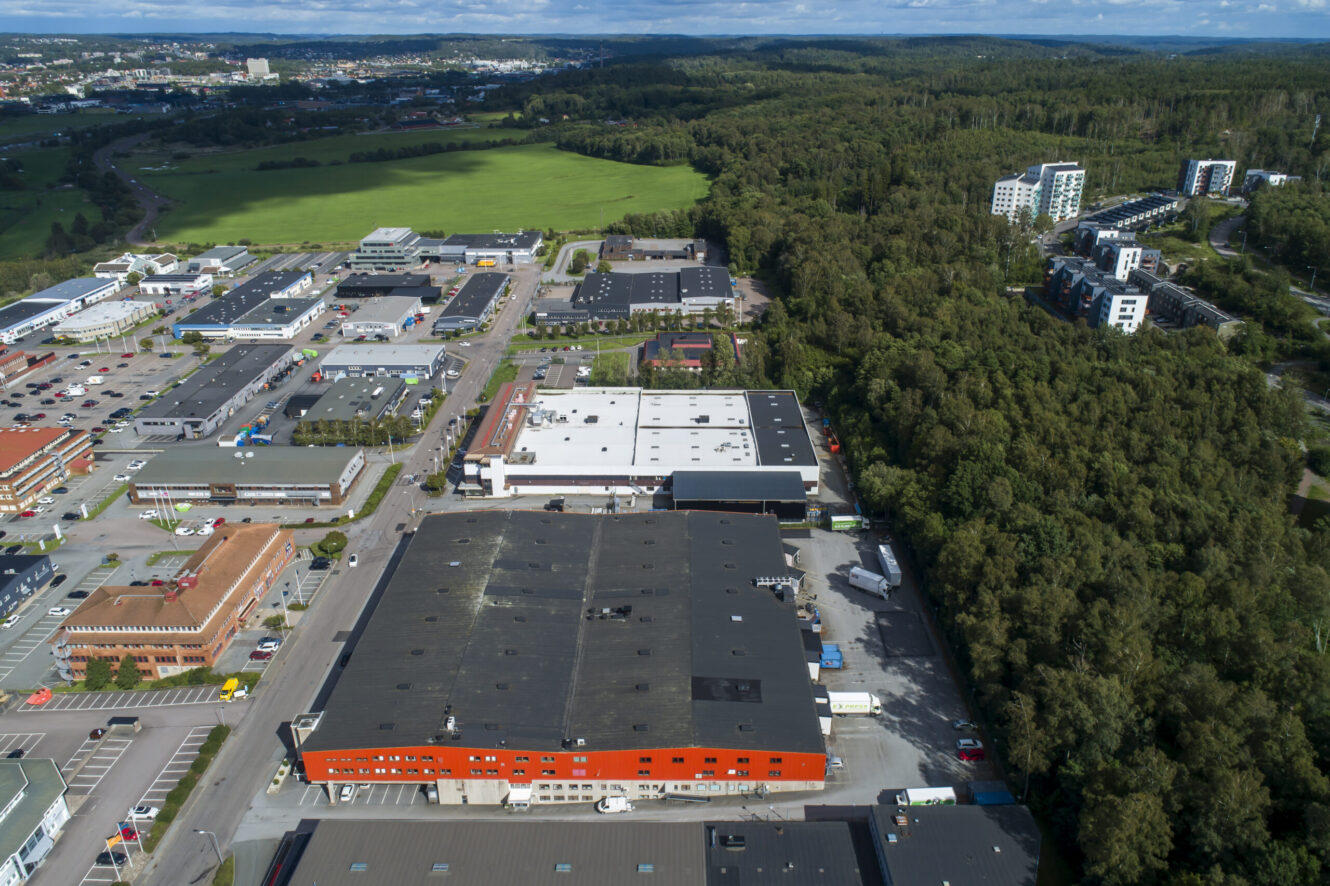 Flygfoto över ett industriområde med stora lagerlokaler intill ett skogsområde, bostadshus och öppna gröna fält i bakgrunden under en delvis molnig himmel.