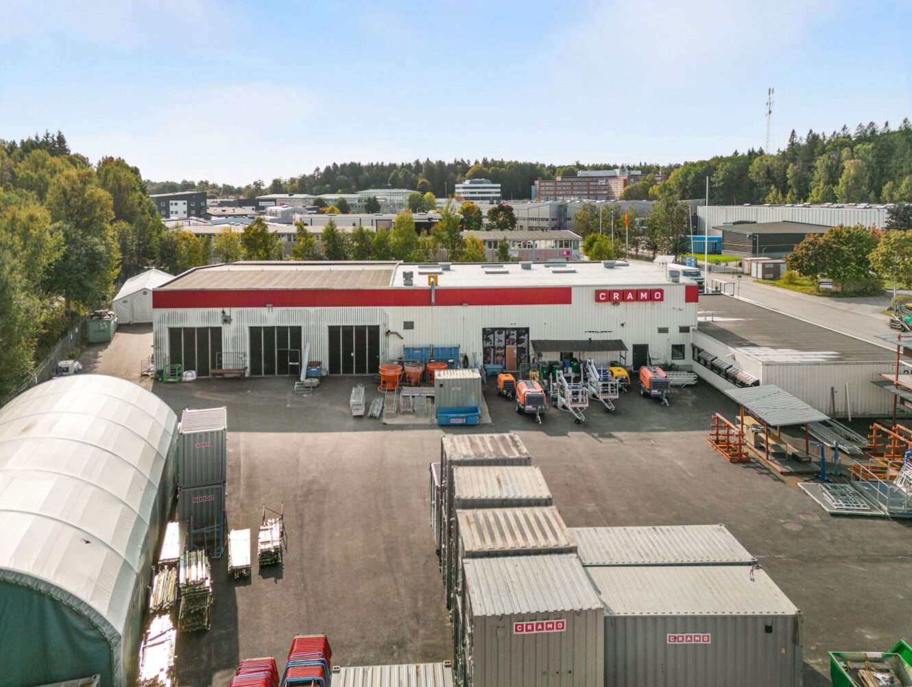 Flygfoto över ett industriområde med en stor vit byggnad märkt Cramo, lagercontainrar, utrustning och träd i bakgrunden.