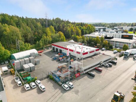 Flygfoto över en kommersiell byggnad med röd dekor, parkeringsplats fylld med bilar, förvaringscontainrar och omgivande träd och industribyggnader.