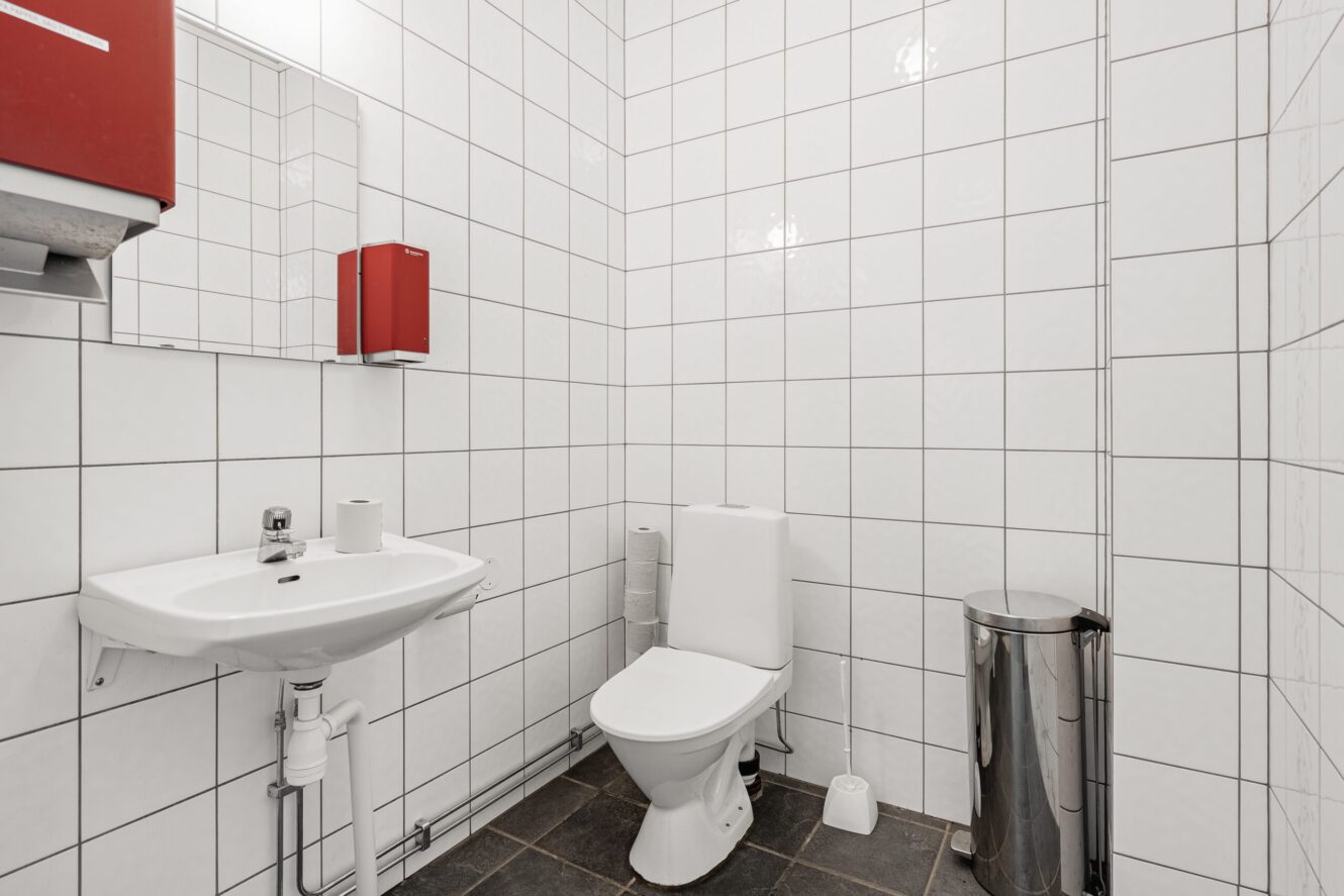 Ett vitkaklat badrum med ett handfat, ett spegelskåp, en toalett, en toalettpappersrulle, en liten papperskorg och en tvålpump på väggen.