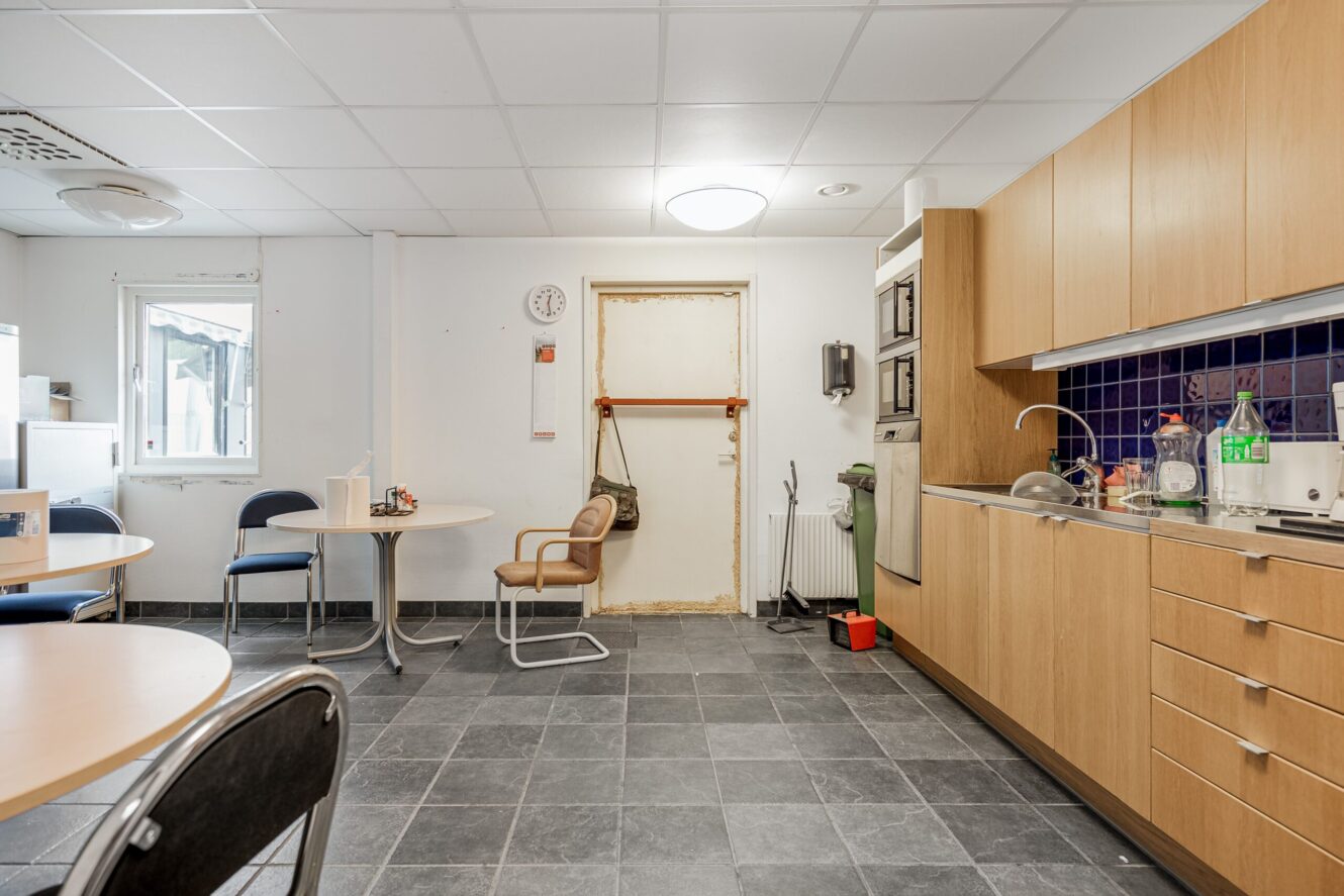 Ett pausrum för personalen med bord, stolar, pentry, väggklocka, mikrovågsugn och rengöringsmedel på ett klinkergolv under fluorescerande lampor.