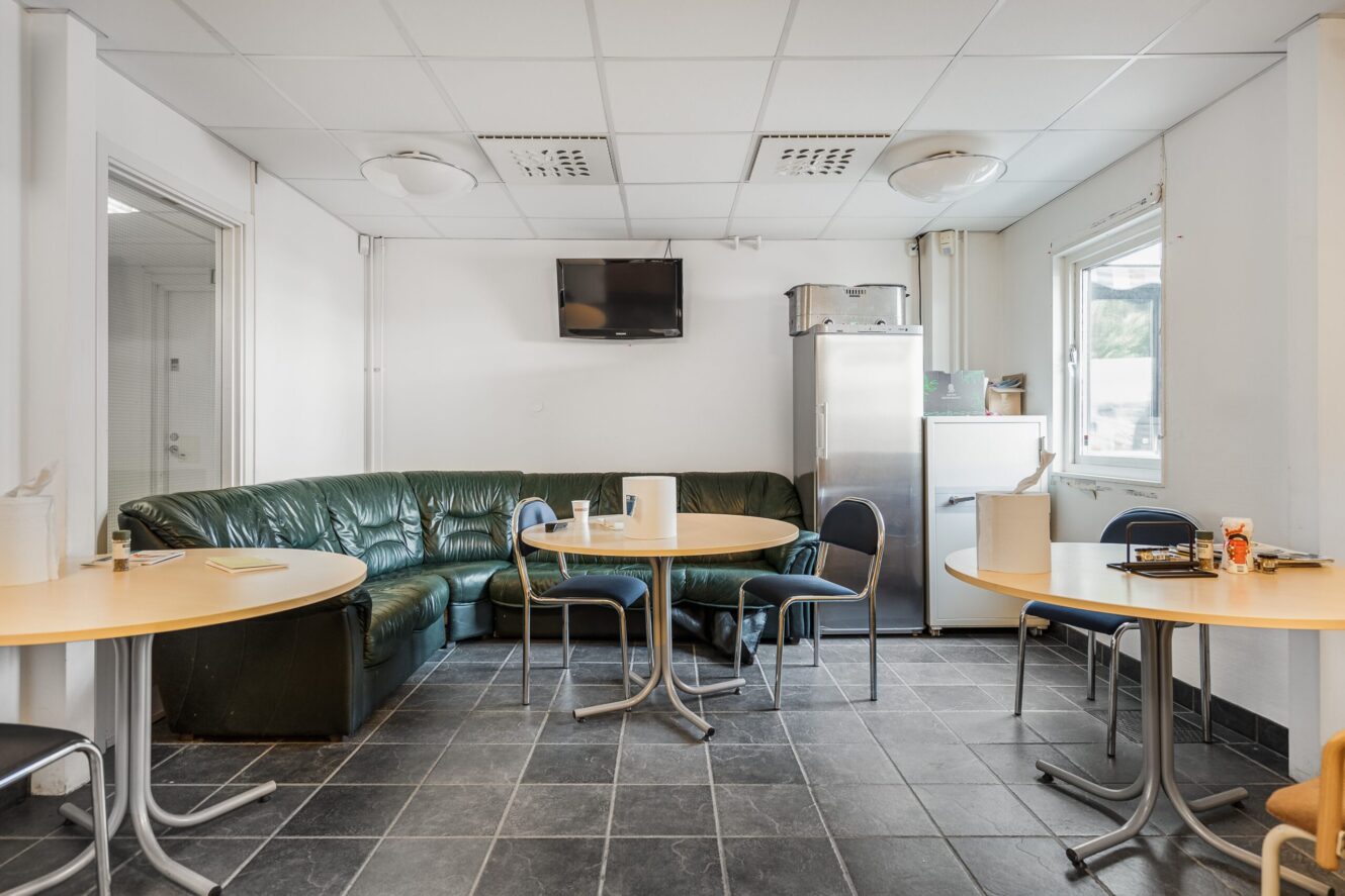 Ett fikarum med en grön bäddsoffa, runda bord med stolar, en väggmonterad TV, kylskåp, mikrovågsugn och pappershanddukar på borden.