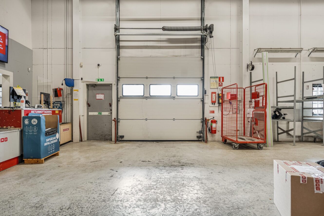 En lagerlokal med en stängd garageport, röda metallställ, hyllor och ett paket på golvet.