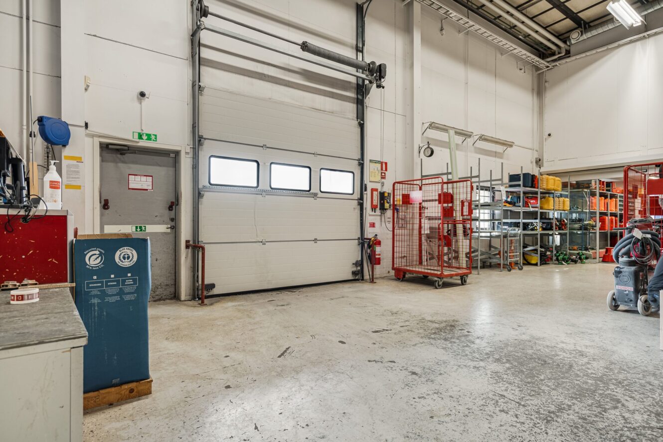 Industriell lagerlokal med stängd takskjutport, metallhyllor, röda burar, säkerhetsutrustning samt olika verktyg och maskiner på ett betonggolv.