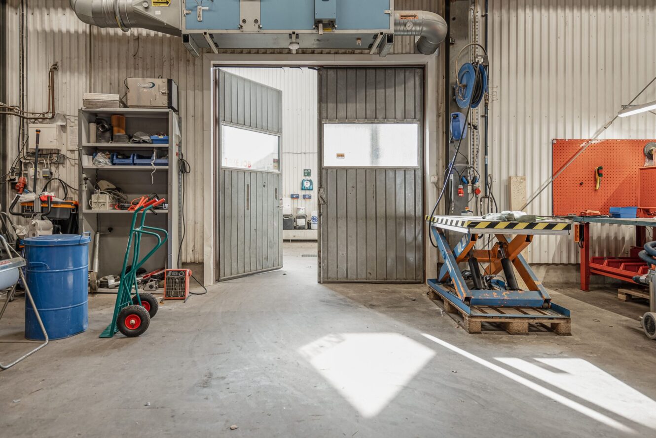 En industriell verkstad med öppna dubbeldörrar, hyllor, verktyg, maskiner, en blå tunna och en handtruck på ett betonggolv.