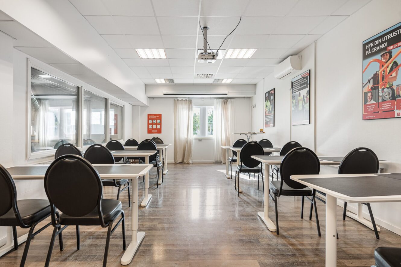 Ett tomt klassrum med flera svarta stolar och vita bord, trägolv, affischer på väggarna och en projektor monterad i taket. Solljuset kommer in genom stora fönster med lätta gardiner.