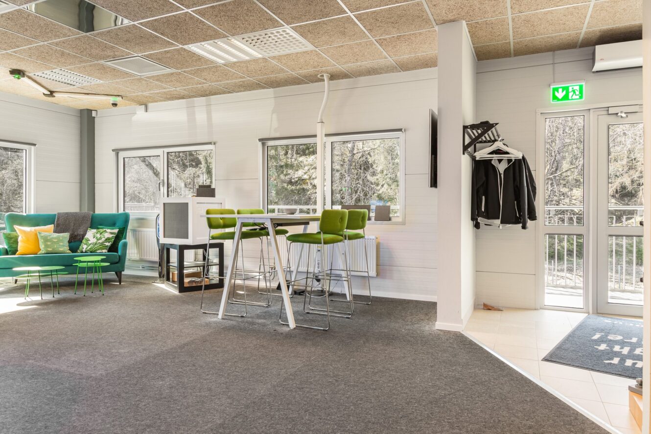 Modern kontorslounge med gröna stolar, soffa med kuddar, klädhängare, höga bord och stora fönster som släpper in naturligt ljus; utgångsdörr till höger.