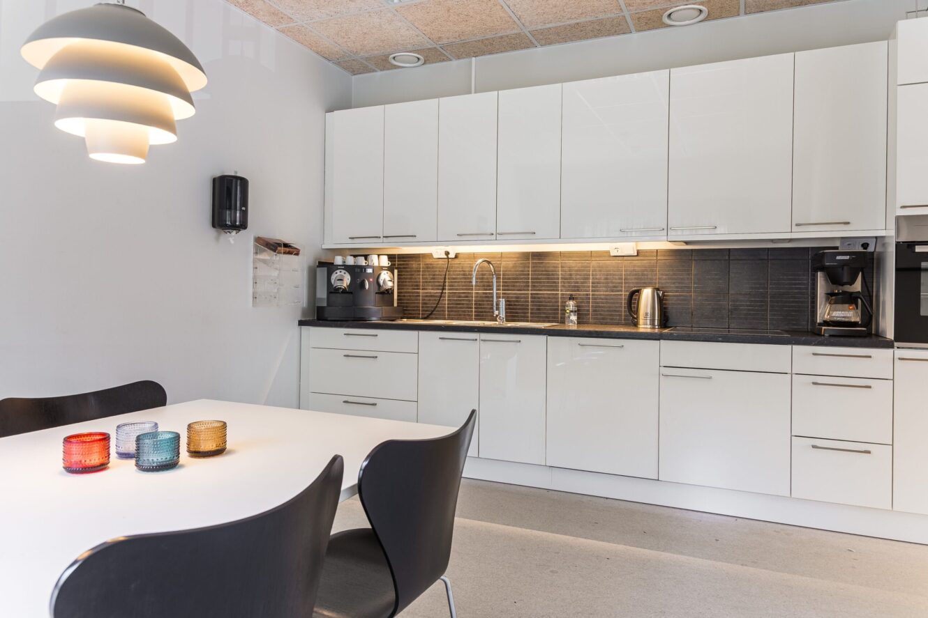 Modernt kök med vita skåp, svart stänkskydd, kaffemaskiner, diskbänk och ett vitt matbord med fyra stolar och färgade ljusstakar.