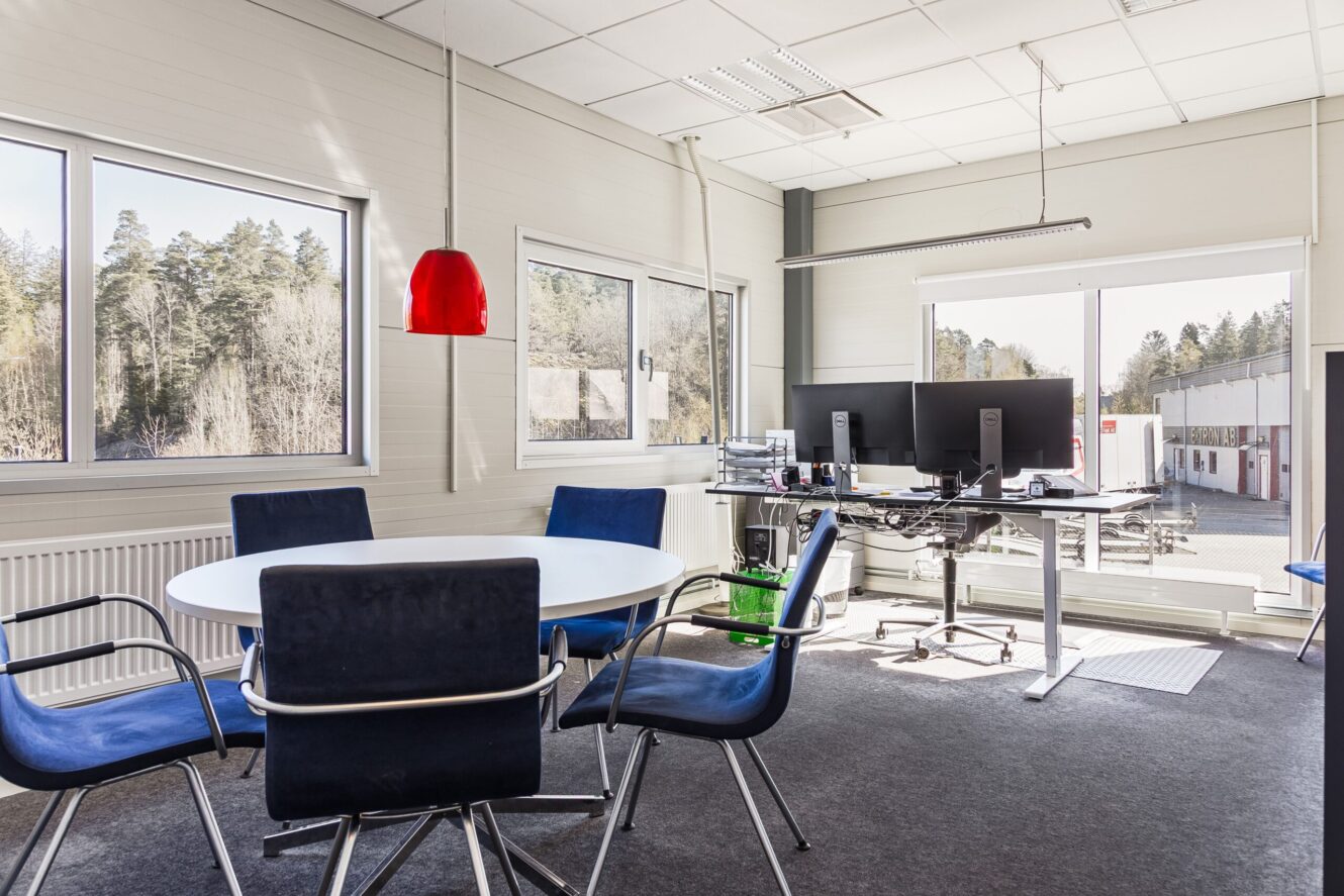 Modernt kontor med ett runt mötesbord, blå stolar, två datorskärmar på ett ståbord och stora fönster med utsikt över träd och byggnader.