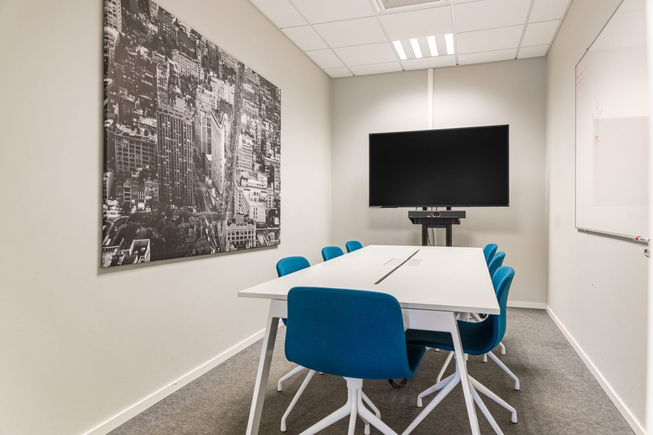 Litet konferensrum med ett vitt bord, sex blå stolar, en väggmonterad TV-skärm, ett stort foto av en stadsbild och en whiteboardtavla.