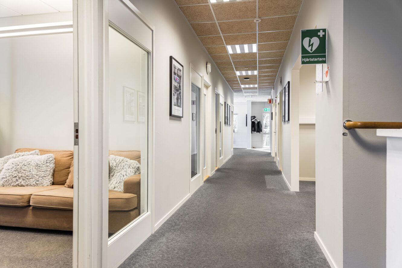 Kontorskorridor med grå heltäckningsmatta, inramade tavlor på väggarna, en brun soffa i ett sidorum och en skylt med en hjärtstartare synlig i taket.