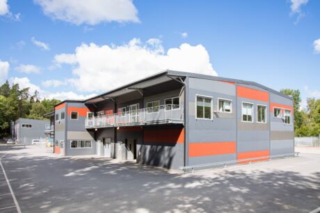 En modern tvåvåningsbyggnad med grå och röd fasadpanel, stora fönster och balkong, belägen på en asfalterad tomt under en delvis molnig himmel.