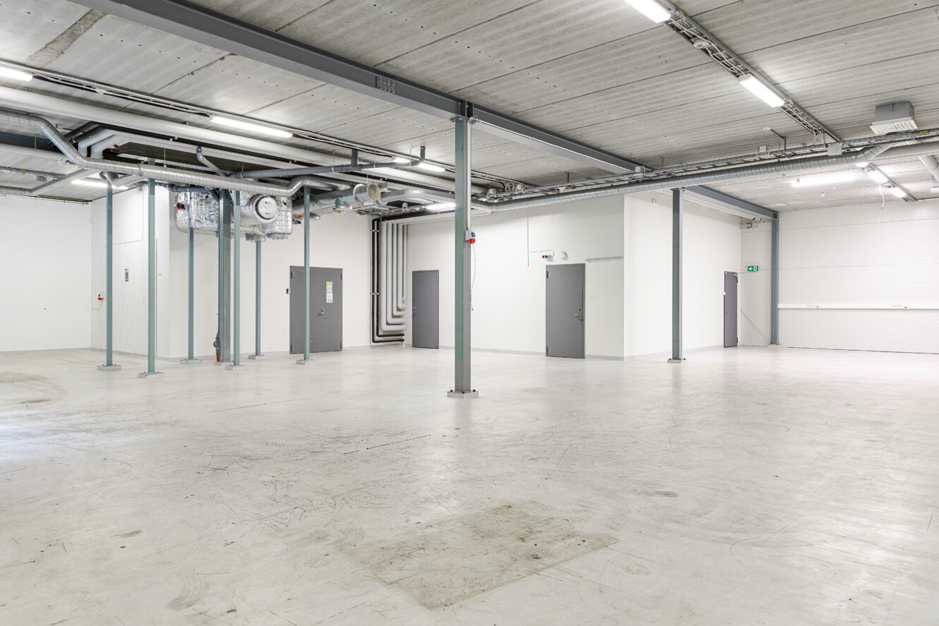 Stor, tom industrilokal med vita väggar, betonggolv, synliga rör i taket, grå dörrar och stålbalkar. Stark takbelysning lyser upp området.