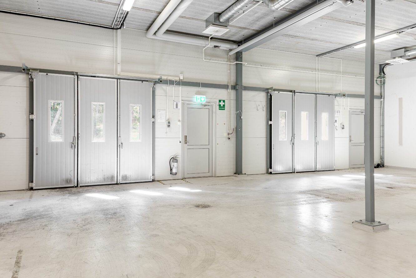 Ett tomt, rent industriellt garage med vita väggar, stora metalldörrar, takbelysning och en branddörr med en grön utgångsskylt.