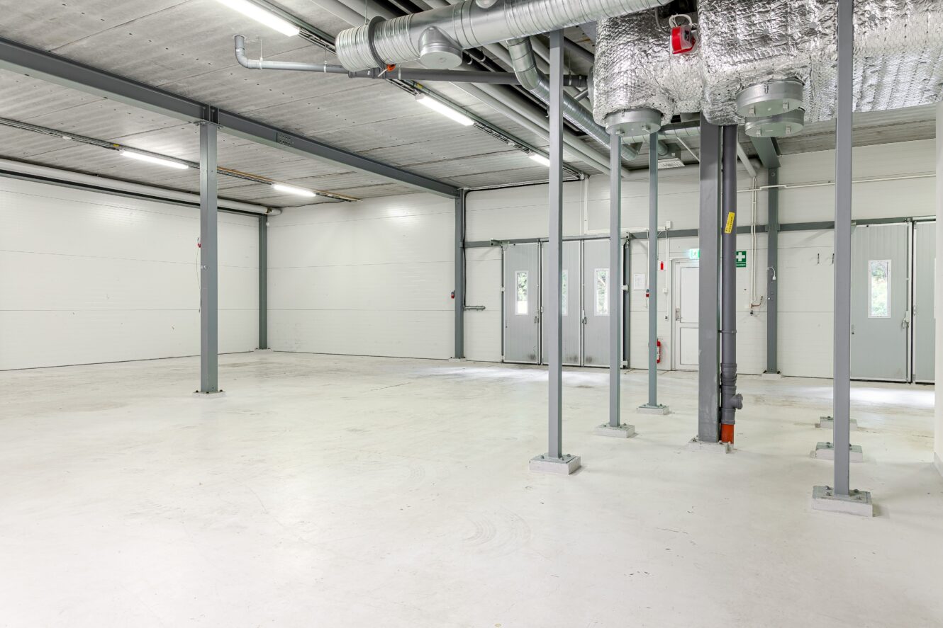 Stor, tom industrilokal med vita golv, metallbalkar, synliga kanaler och flera dörrar längs bakväggen.
