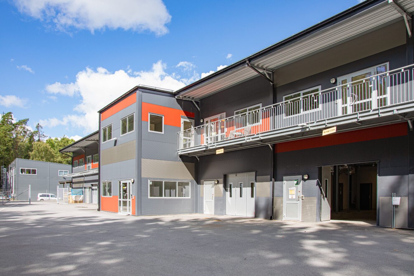 Modern affärs- eller industribyggnad med grå och orange fasad, stora fönster, balkonger på övre plan och flera ingångar, under en delvis molnig himmel.