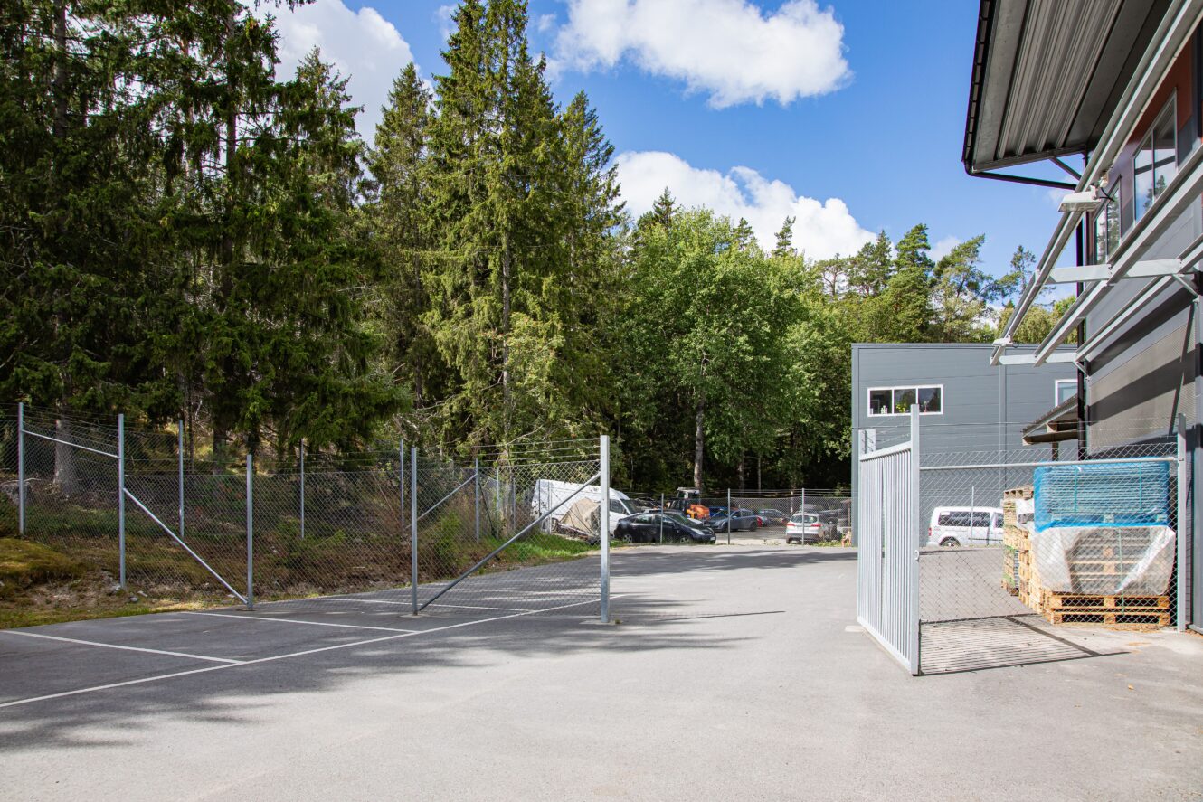 En inhägnad industritomt med öppna grindar, staplade material på pallar och flera bilar parkerade nära träd i bakgrunden under en delvis molnig himmel.