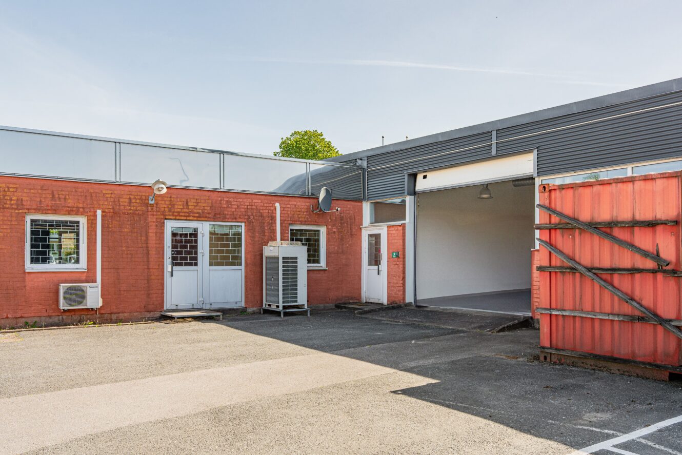 En industribyggnad i rött tegel med gallerförsedda fönster, stängda och öppna dörrar, en utomhusenhet och ett öppet garage som leder till en tom interiör.