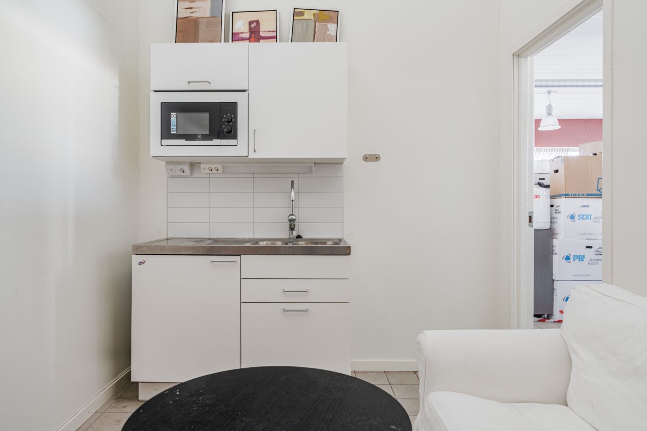 Litet vitt pentry med diskho, mikrovågsugn och skåp; i förgrunden ett svart runt bord och en vit fåtölj. Ovanpå skåpen står konstramar.