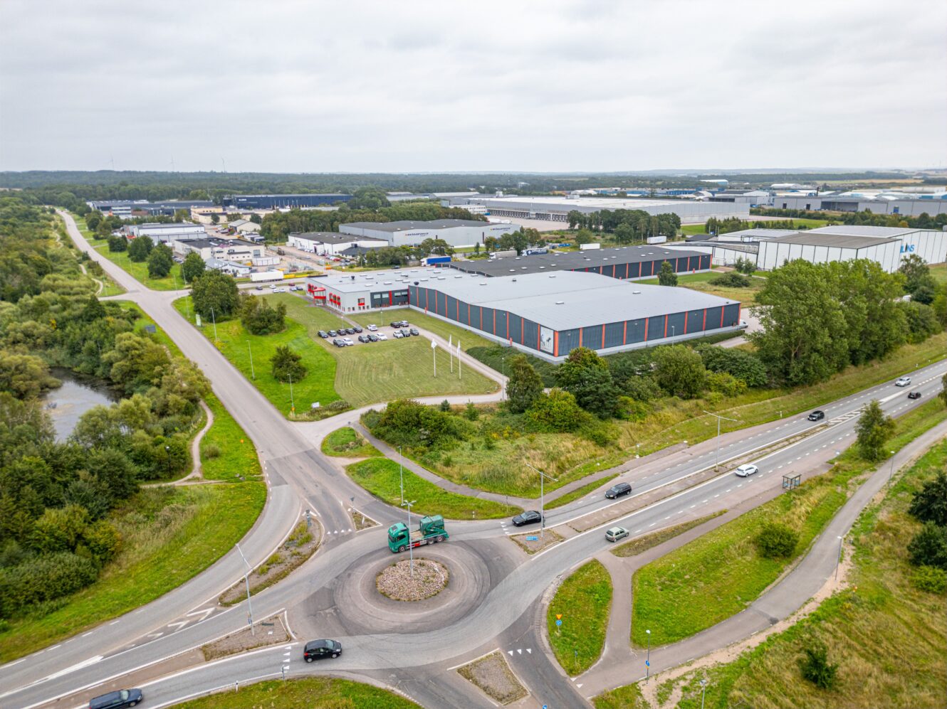 Flygfoto över en rondell med flera vägar, omgiven av grönområden och industrihallar under en molnig himmel.