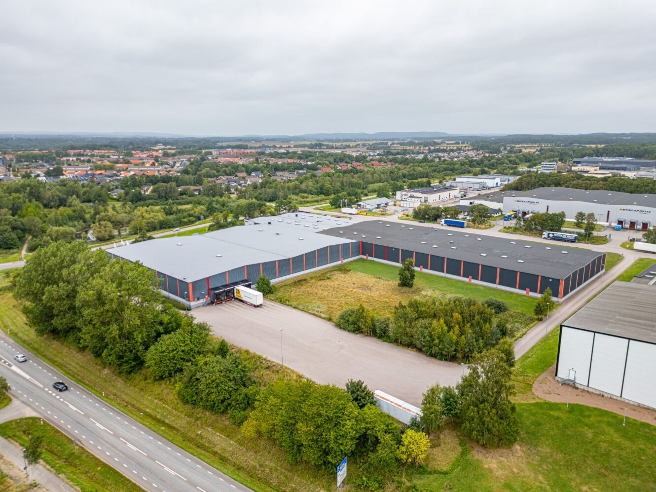 Flygfoto över ett stort industrilager med lastkajer och parkerade lastbilar, omgivet av grönska och intilliggande vägar i ett förortsområde.