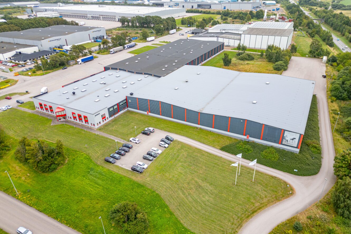 Flygfoto över ett stort industrilager med en parkeringsplats och flera parkerade bilar, omgivet av grönt gräs och närliggande vägar.