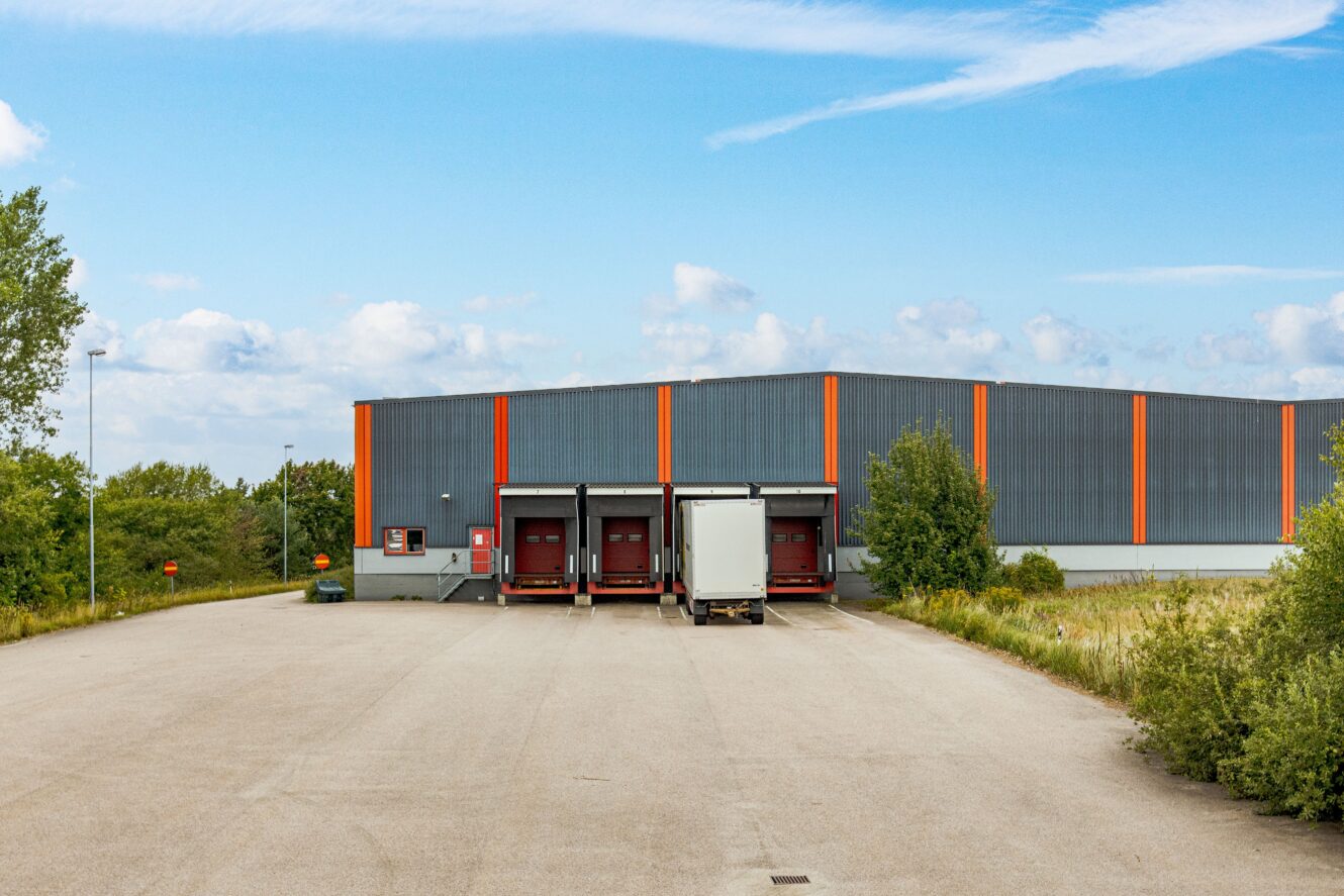 En lagerlokal med tre lastkajer, varav en är upptagen av en lastbil, omges av grönska under en delvis molnig himmel.