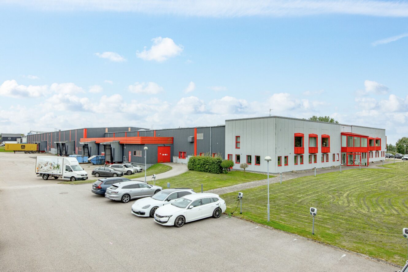 En stor industribyggnad med röda accenter, flera parkerade bilar, gräsytor och en delvis molnig himmel.