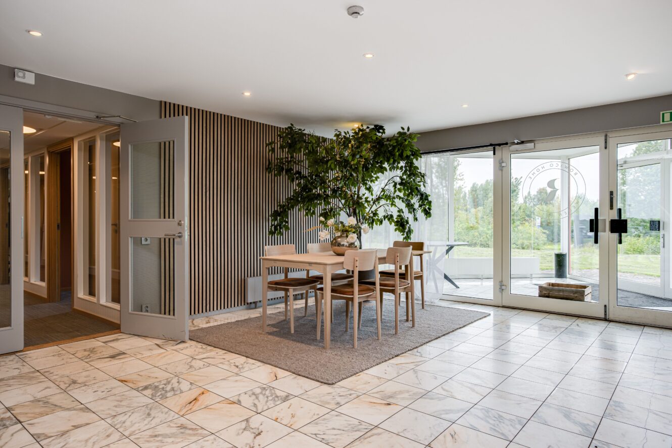 Ett modernt, väl upplyst rum med marmorgolv, ett ljust träbord med sex stolar, en stor inomhusväxt och glasdörrar som leder ut.