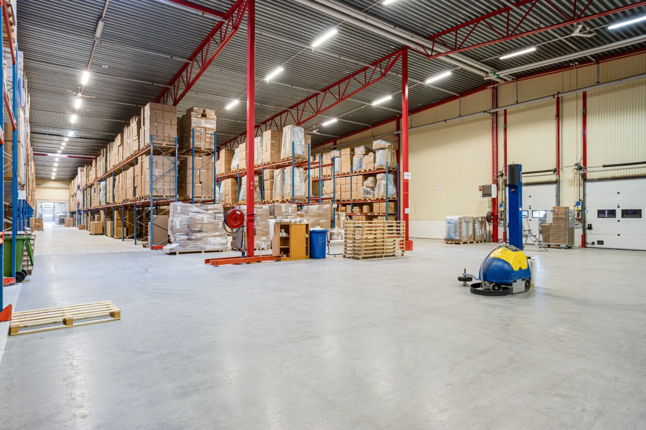 Stor lagerlokal med höga hyllor fulla av lådor och pallar, industriell rengöringsmaskin på golvet och högt i tak med röda stödbalkar.