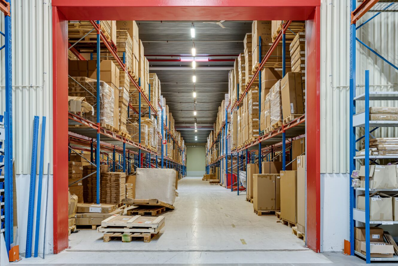 Lagerlokal med höga hyllor staplade med kartonger och lastpallar, sett genom en bred entré med röd ram.