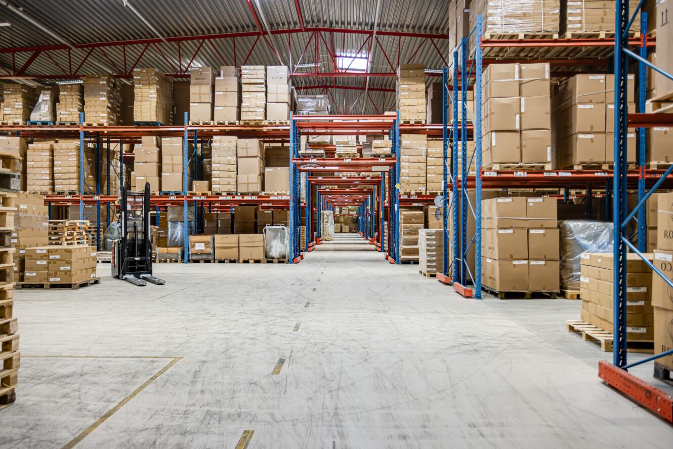 En stor lagerlokal med höga hyllor staplade med kartonger och pallar, med en gaffeltruck till vänster och breda gångar mellan hyllorna.