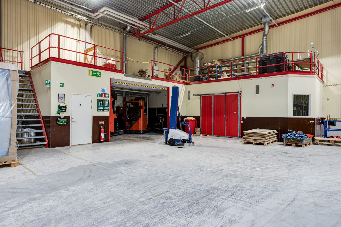 Ett rymligt industrilager med röda räcken, maskiner, förvaringspallar, säkerhetsskyltar och ett entresolplan. Golvet är mestadels fritt med lite utrustning placerad runt omkring.