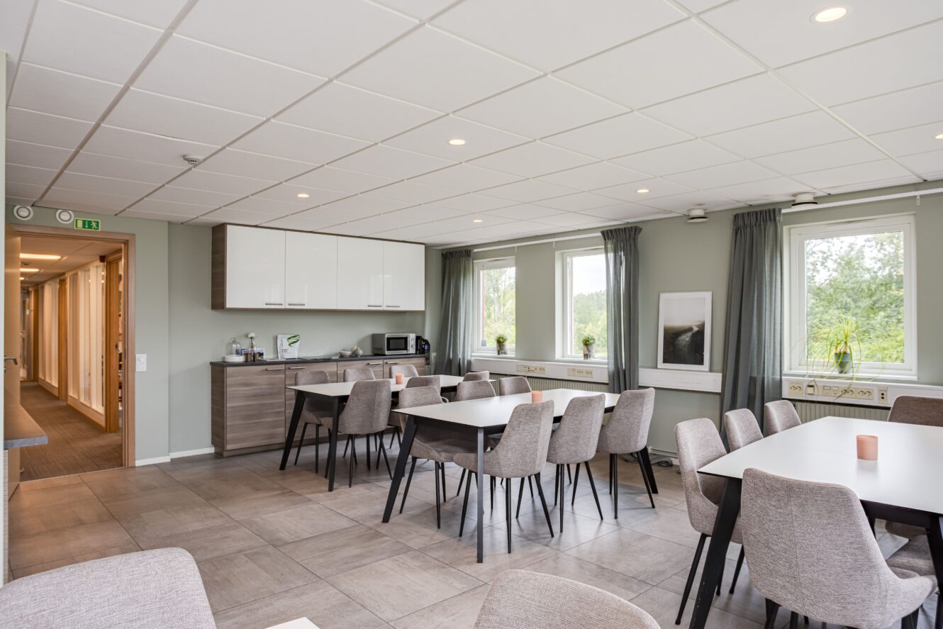 Ett modernt fikarum med flera bord och stolar, ett pentry med skåp och vitvaror, stora fönster och ljusgrå väggar.