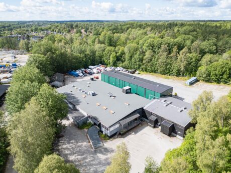Flygfoto över en industrianläggning med flera byggnader, parkeringsplatser och omgivande träd, belägen nära ett skogsområde och ett avlägset bostadsområde.