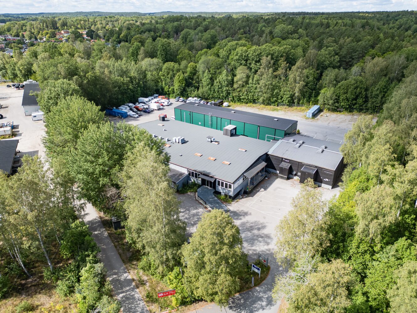 Flygfoto över en stor industribyggnad med grönt tak, omgiven av träd och parkeringsplatser, belägen i utkanten av ett skogsområde.