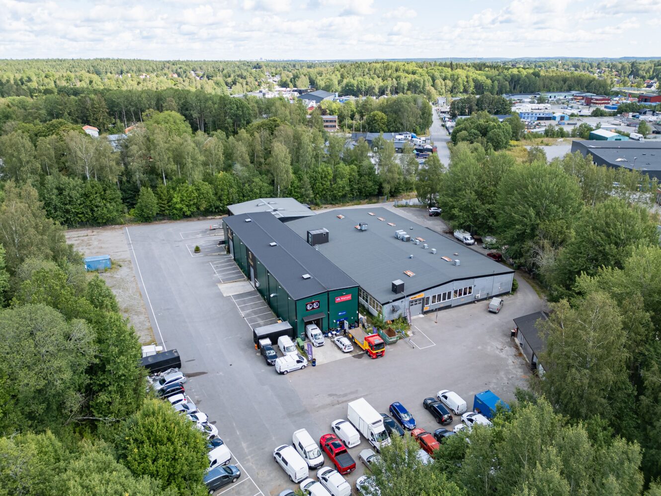 Flygfoto över en stor kommersiell byggnad med parkerade fordon framför, omgiven av träd och parkeringsplatser, med fler byggnader och grönska i bakgrunden.