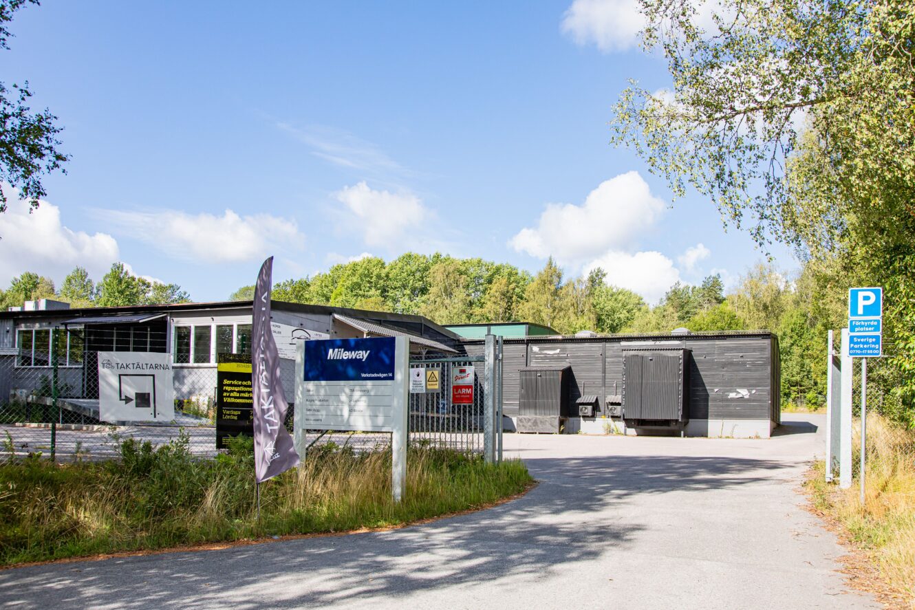 Industrianläggning med en grindförsedd entré, skyltar, parkeringsplats och träd i bakgrunden en solig dag.