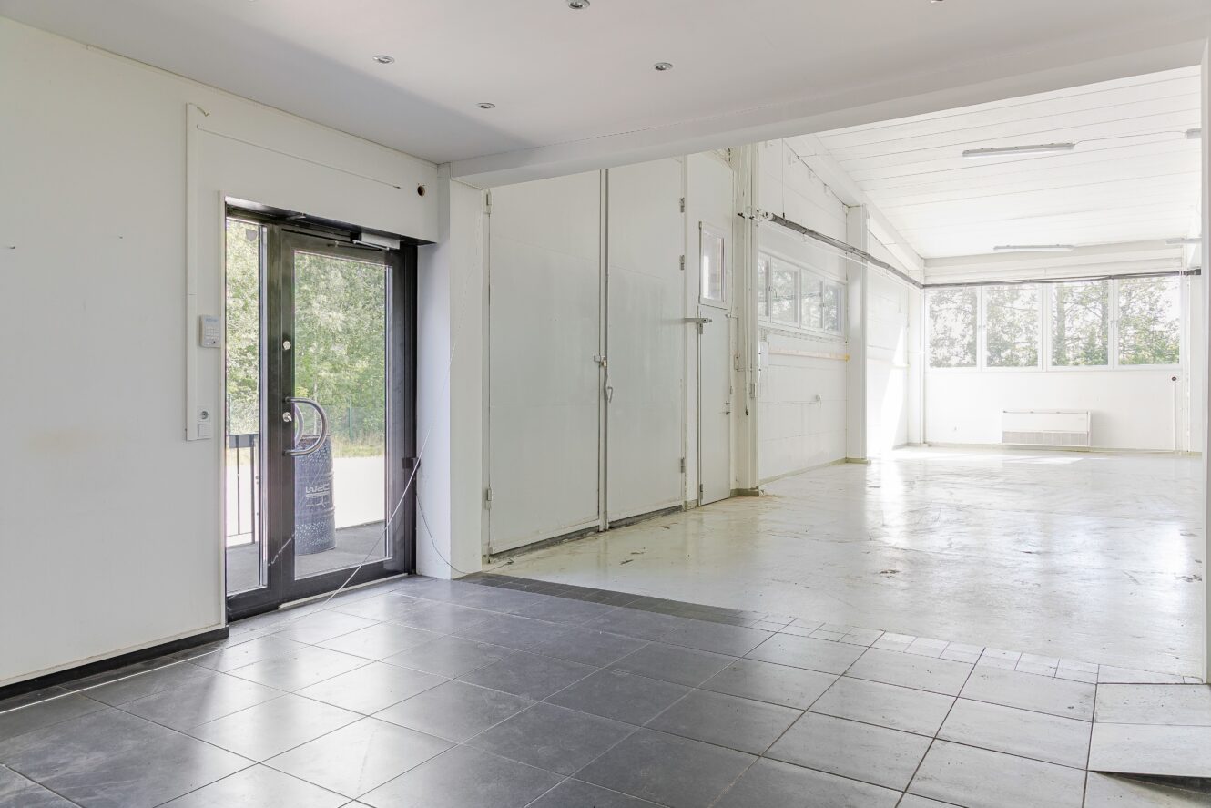 En ren, tom industrilokal med klinker- och betonggolv, stora fönster, vita väggar och en glasdörr som leder ut.