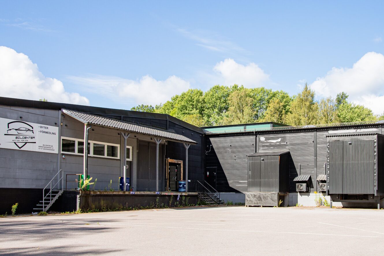 En modern industribyggnad med lastkajer, metalltrappor och en skylt på utsidan, omgiven av träd under en delvis molnig himmel.