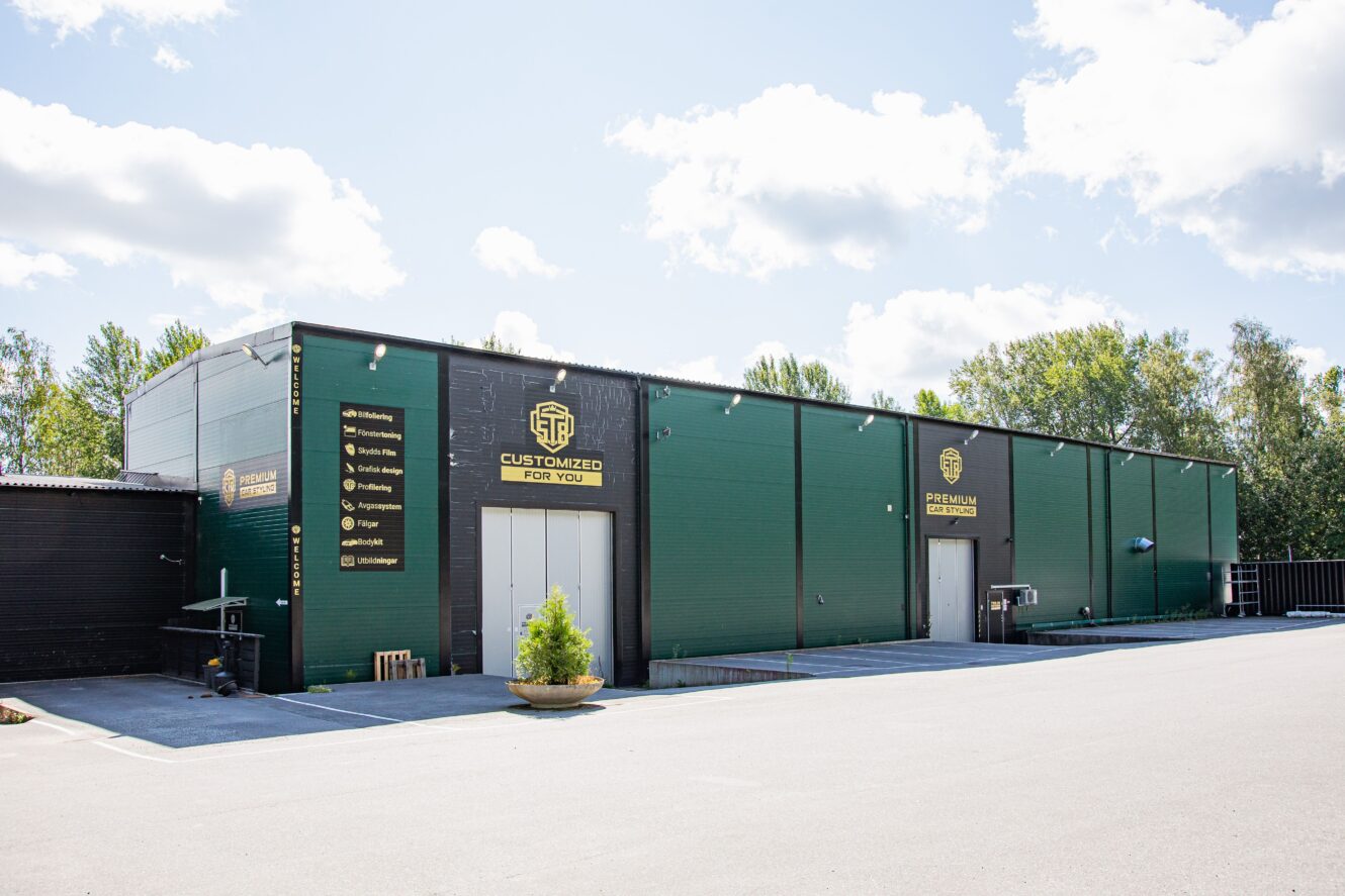 Stor grön industribyggnad med företagslogotyper och skyltar på utsidan, mot en bakgrund av träd och en delvis molnig himmel.